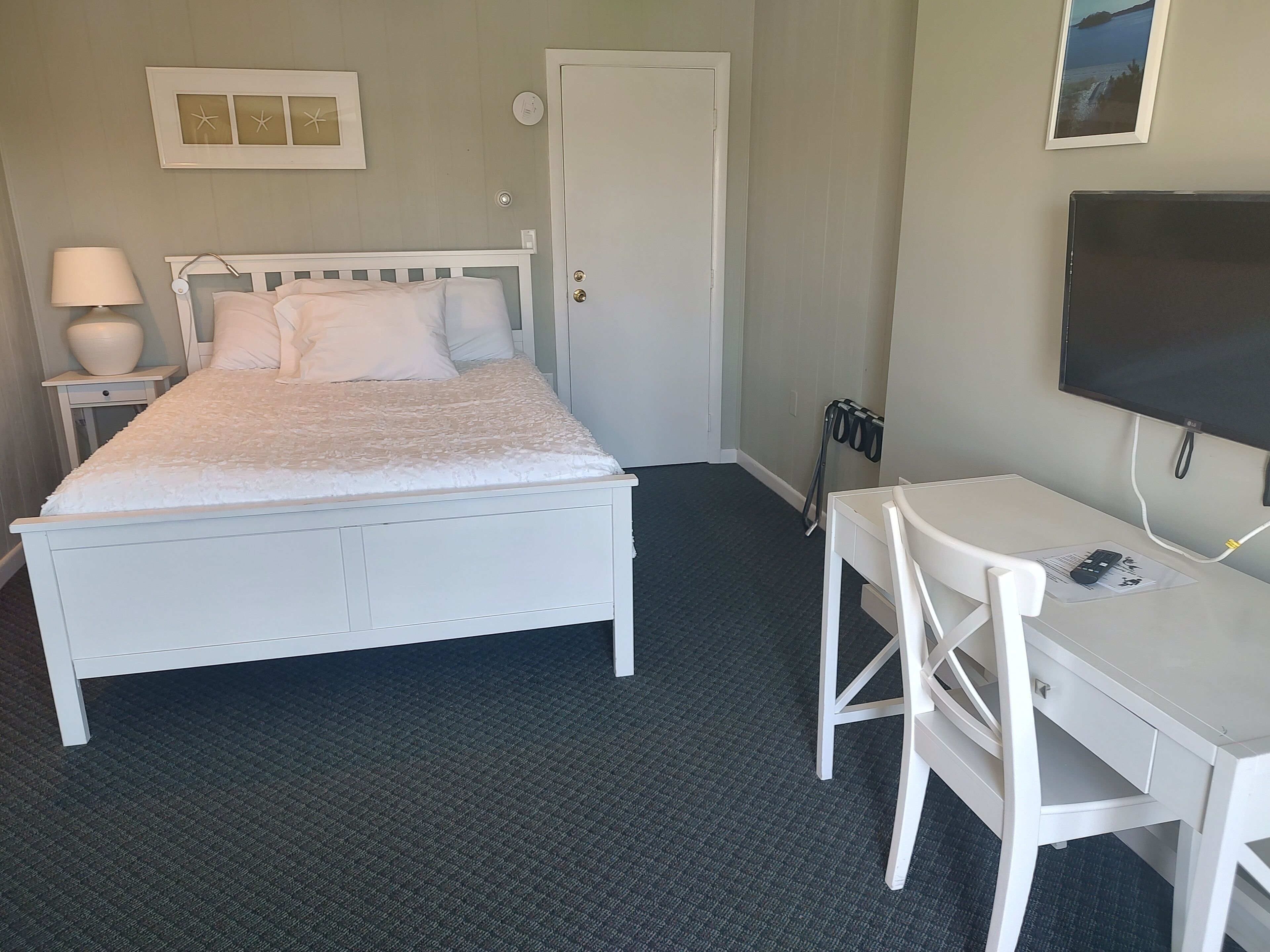 A bedroom with a bed, bedside table with a lamp, a desk with a chair, a TV, and a door.
