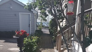 A paved pathway leading to a white building with a door and windows. There is a potted plant with red flowers and a wooden fence with lattice panels.