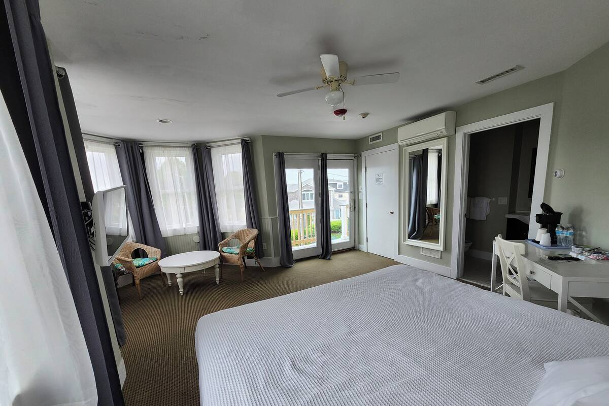 A hotel room with a large bed, a desk with a mirror, a chair, a small table, and a ceiling fan.