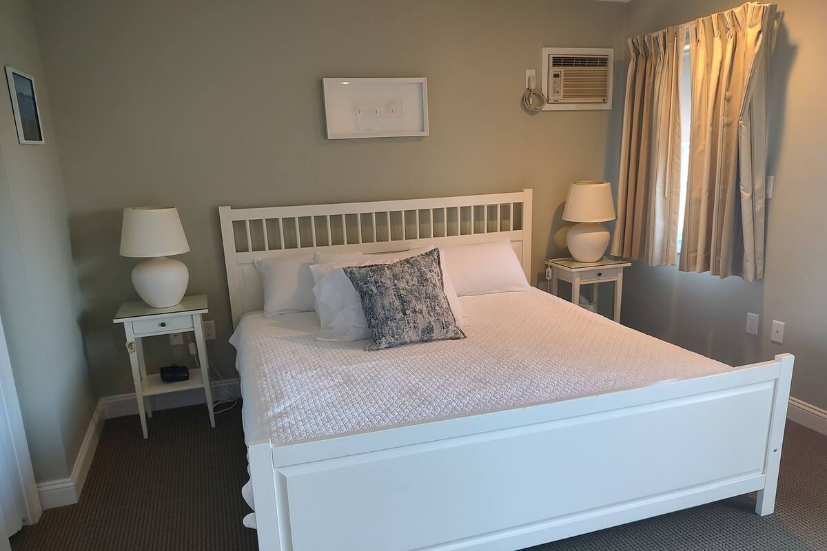 A neatly arranged bedroom with a white bed, two bedside tables with lamps, a wall-mounted air conditioning unit, and a window with curtains.