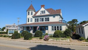 A large, two-story house with a prominent tower, a porch with American flags, and a well-maintained front yard with various shrubs and flowers.