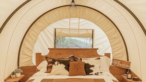 A cozy tented room with a wooden bed, a small table with a lamp, and a sign that says "enjoy the little things."