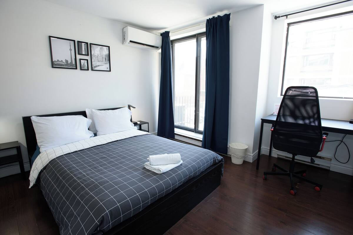 A neatly made bed with a checkered bedspread, a desk with a chair, and a window with curtains.