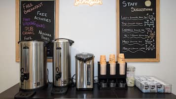 Una estación de café con dos máquinas de café, una pila de vasos desechables y una pizarra con indicaciones sobre el desayuno gratuito y sugerencias del personal.