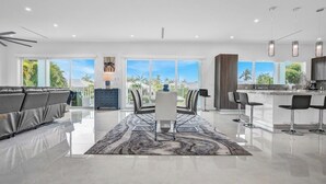 A modern living room with a sofa, a dining table with chairs, a kitchen area with a bar, and large windows offering a view of palm trees. - P70 - Stunning 5 bed, 5 bath home with dockage and oversized private pool in Key Colony! (Key Colony Beach)