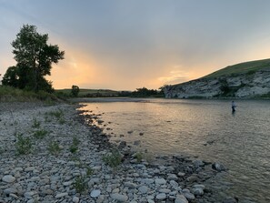 A riverbank with a rocky foreground, a lone tree, and a person fishing.