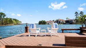 Una terraza de madera con dos sillas blancas y una pequeña mesa, con vista a un lago y casas al fondo.