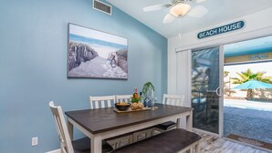 A dining area with a dark wooden table, white chairs, and a bench. A painting of a beach path hangs on the wall.