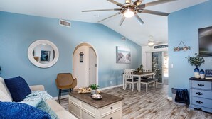 A living room with a ceiling fan, a round mirror, a dining area with a table and chairs, and a kitchen area with a TV and cabinets.