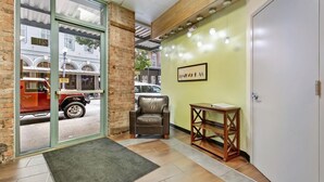 Un hall d’hôtel avec un mur en briques, un plafond en bois, un mur végétal agrémenté d’un tableau encadré, un fauteuil en cuir, une petite table en bois et une porte avec un panneau indiquant la sortie.