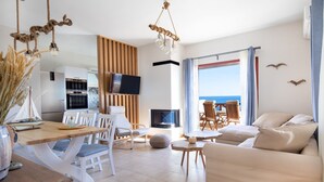 Un salon moderne avec un canapé, une table à manger et des chaises. Un téléviseur est fixé au mur et il y a une cheminée. La pièce est dotée d’une grande fenêtre offrant une vue sur la mer.