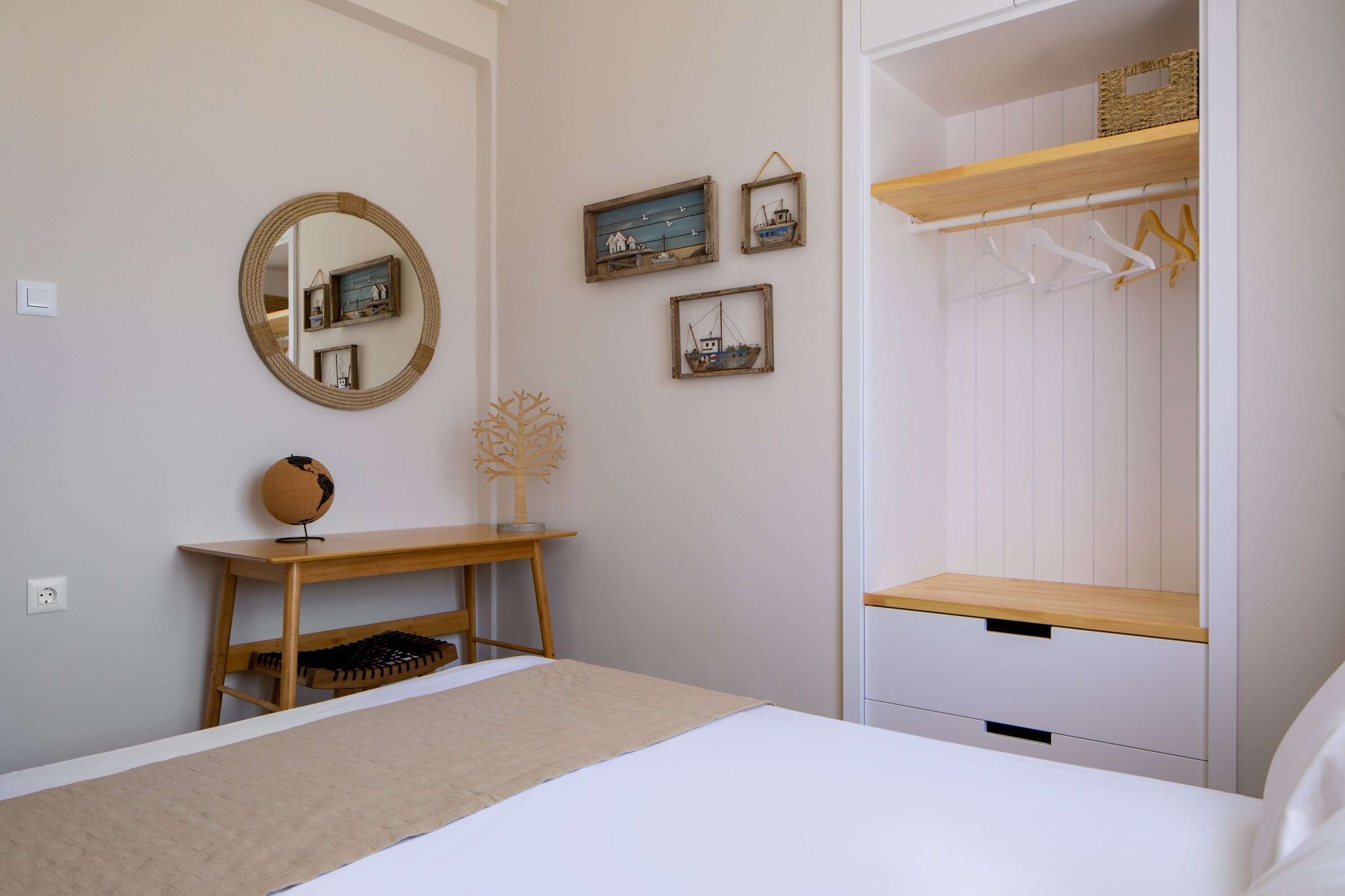 A bedroom with a bed, a wooden desk, a mirror, and a wardrobe with hanging clothes.