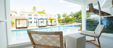 A poolside area with wicker chairs and a glass balustrade overlooking a pool and a building with multiple windows.