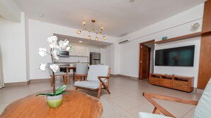 A modern living room with a wooden dining table, two chairs, a TV, and a kitchen in the background. - 1 Bedroom Garden View Presidential Suites Cabarete (Sosúa)