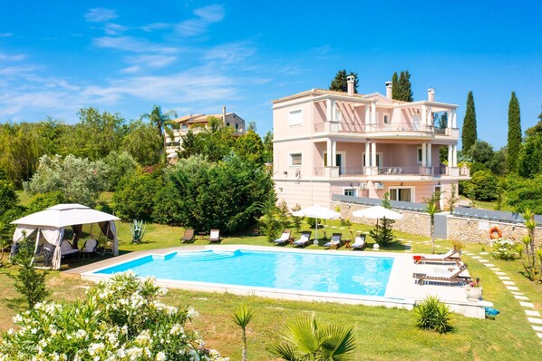 A multi-story building with a swimming pool, surrounded by lush greenery and a clear sky.