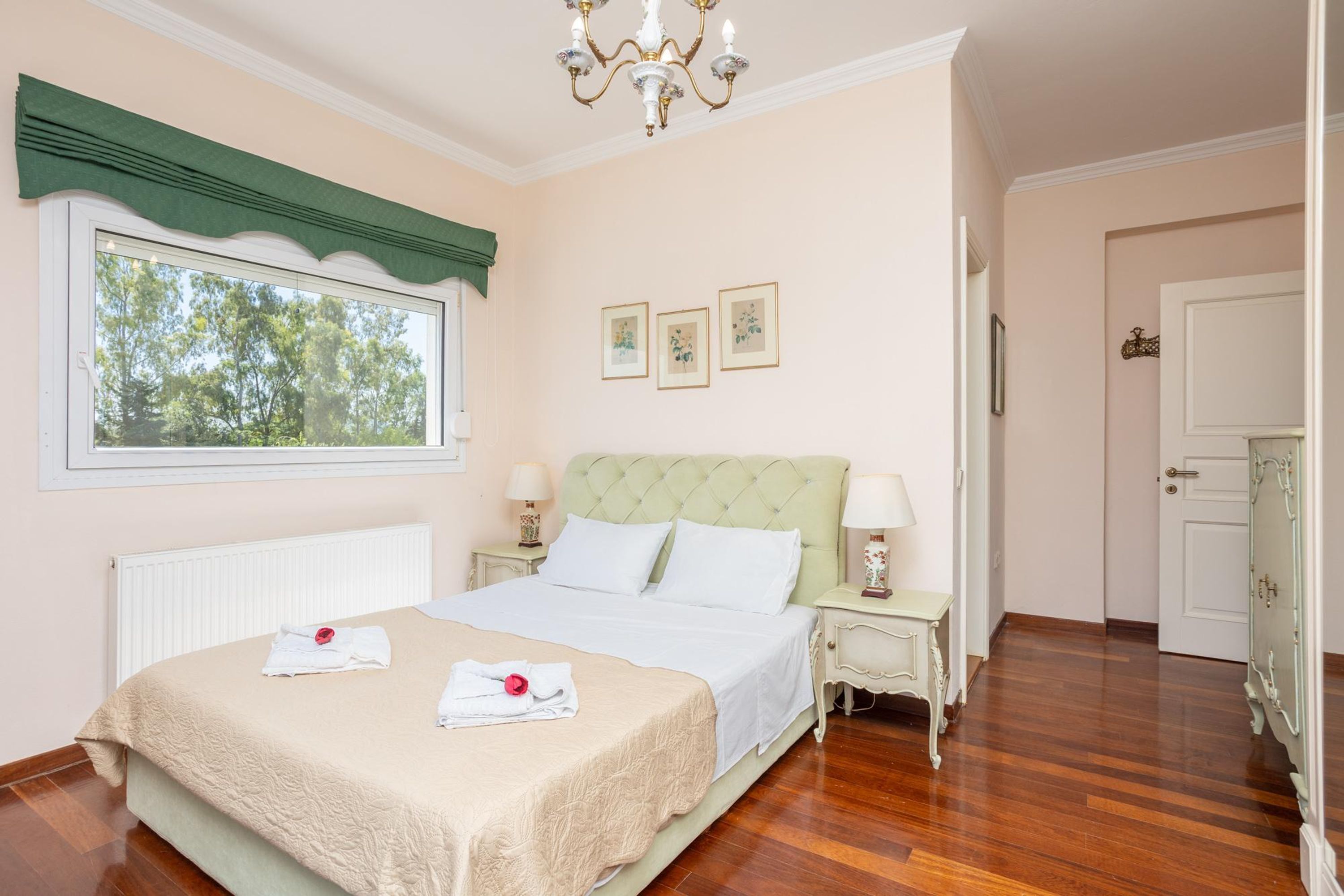 Une chambre à coucher bien aménagée, avec un grand lit, deux tables de chevet avec des lampes, une fenêtre agrémentée d’un lambrequin vert et un lustre.