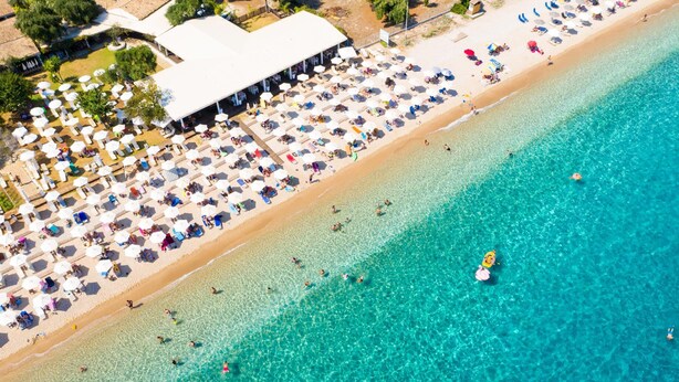 An aerial view of a beach with numerous umbrellas and sun loungers, a clear blue sea, and a building with a white roof. - Villa Denise in Corfu (Corfu)