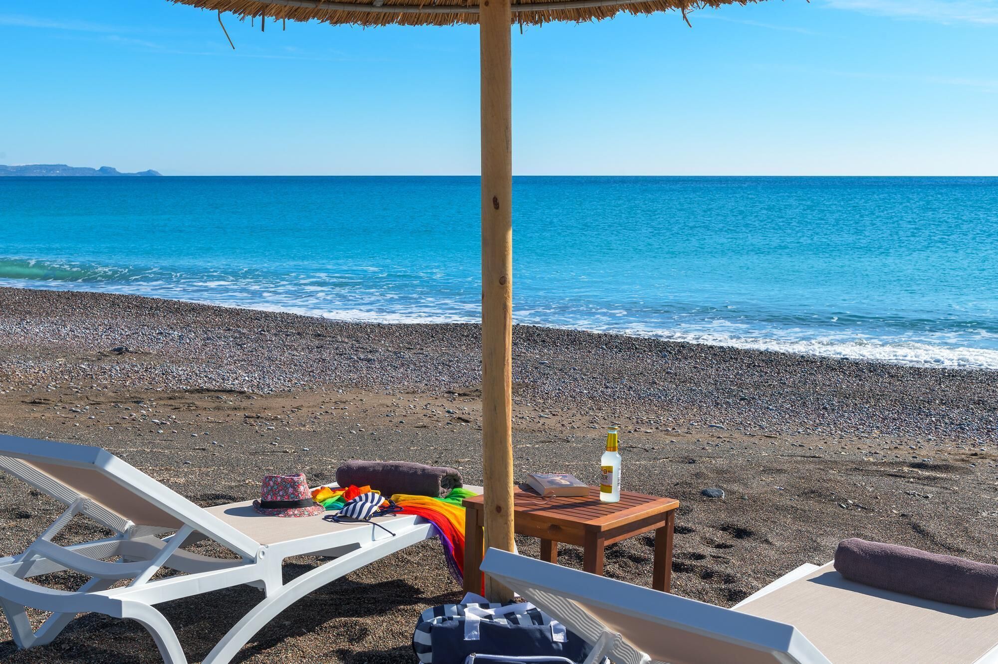 A beach with two white lounge chairs, a table with a bottle, a hat, and a colorful umbrella.