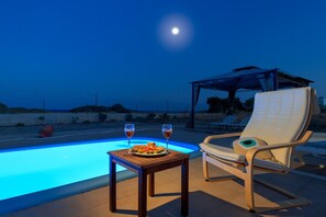 Escena junto a la piscina con una mesa y dos sillas, un área de descanso cubierta y vista a la playa por la noche.