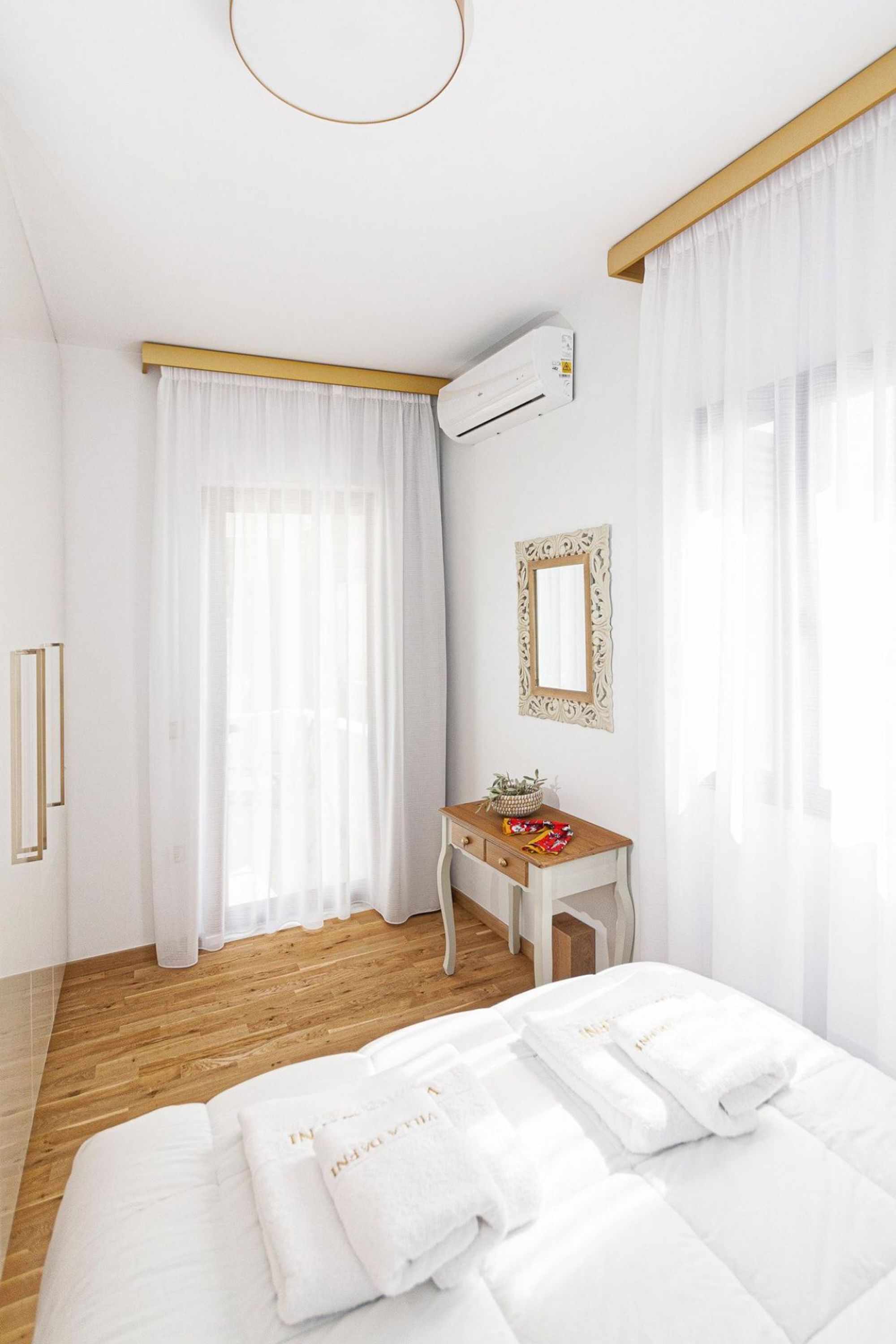 Une chambre avec un plancher en bois, un lit avec une literie blanche, un bureau avec un miroir et un appareil de climatisation.