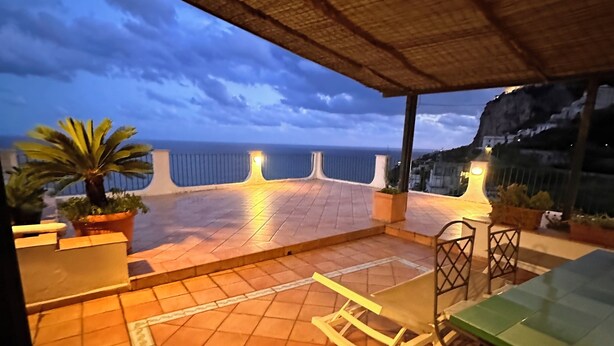 An outdoor patio with a tiled floor, a thatched roof, a table and chairs, and a view of the sea.