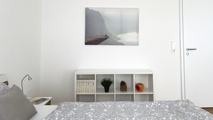 A bedroom with a bed, a white wall, a white door, a white shelf with books and decorative items, and a framed picture of a landscape. - Charming & central living in Weiden - Hej (Weiden in der Oberpfalz)