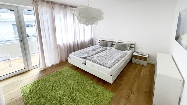 A modern bedroom with a large bed, a green rug, a white dresser, and a balcony with curtains. - Charming & central living in Weiden - Hej (Weiden in der Oberpfalz)