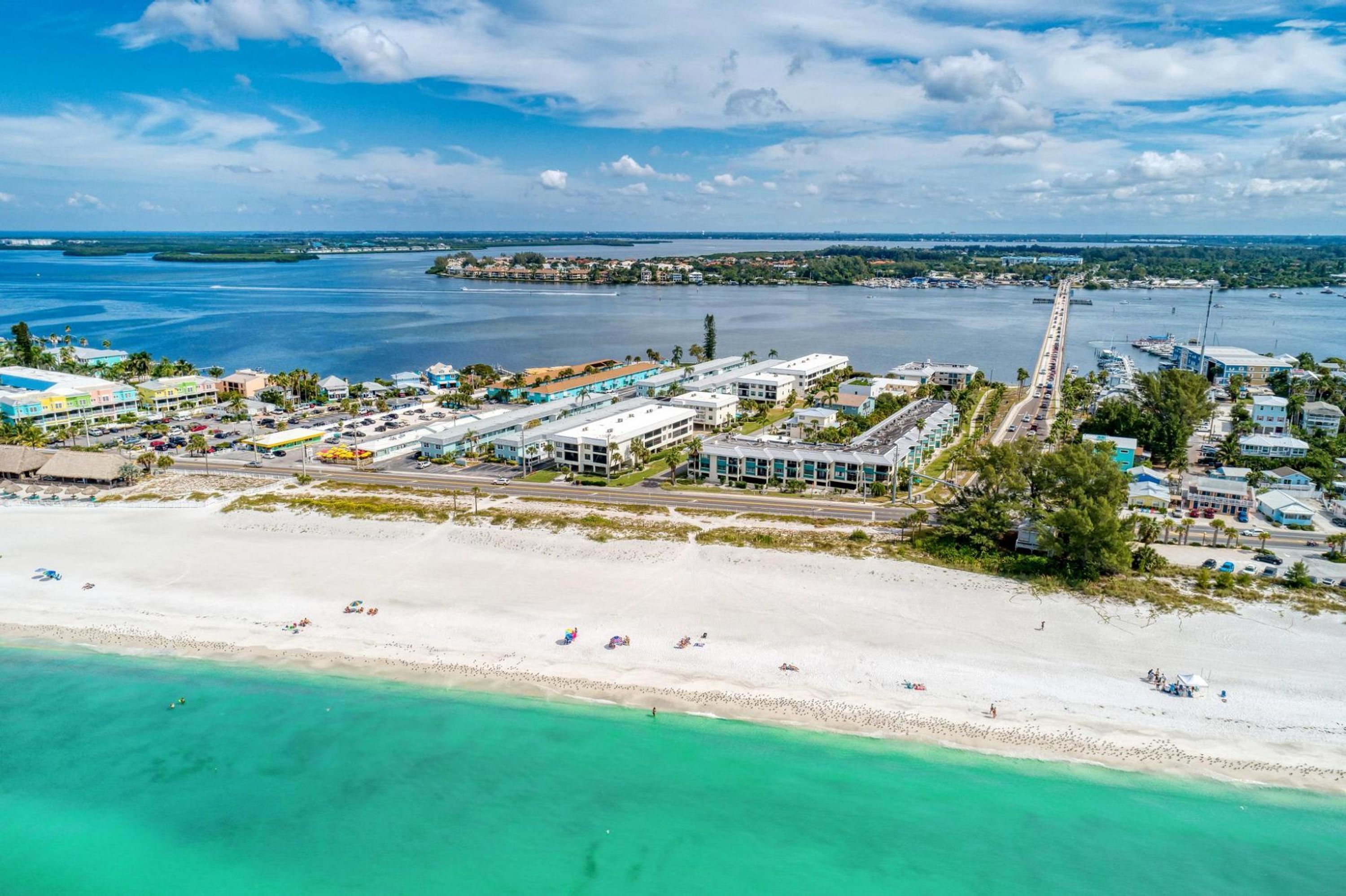 An aerial view of a coastal area with a beach, a marina, and a row of buildings with a long pier extending into the water.