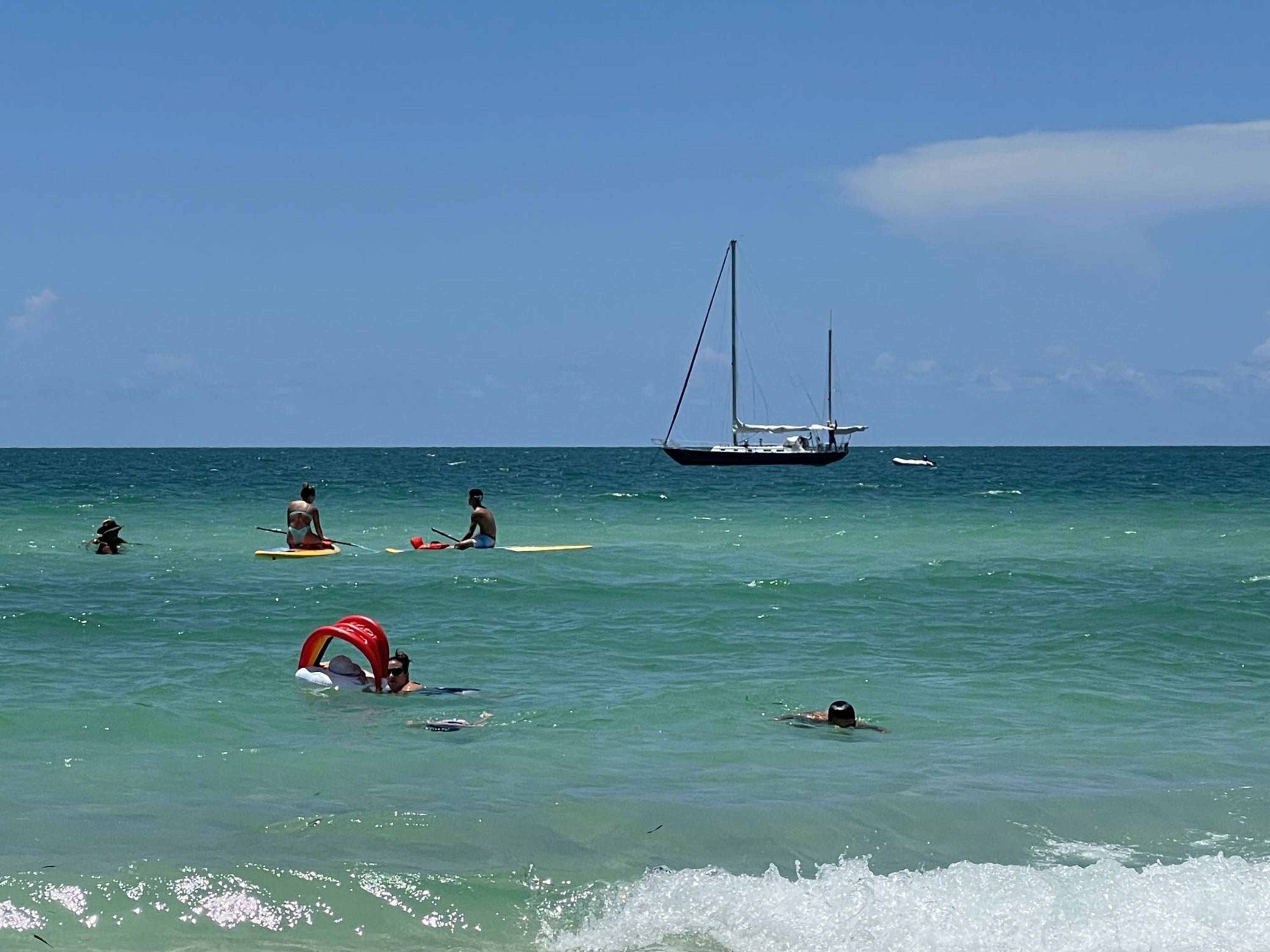 A beach with people engaging in water activities, a sailboat, and a clear sky.