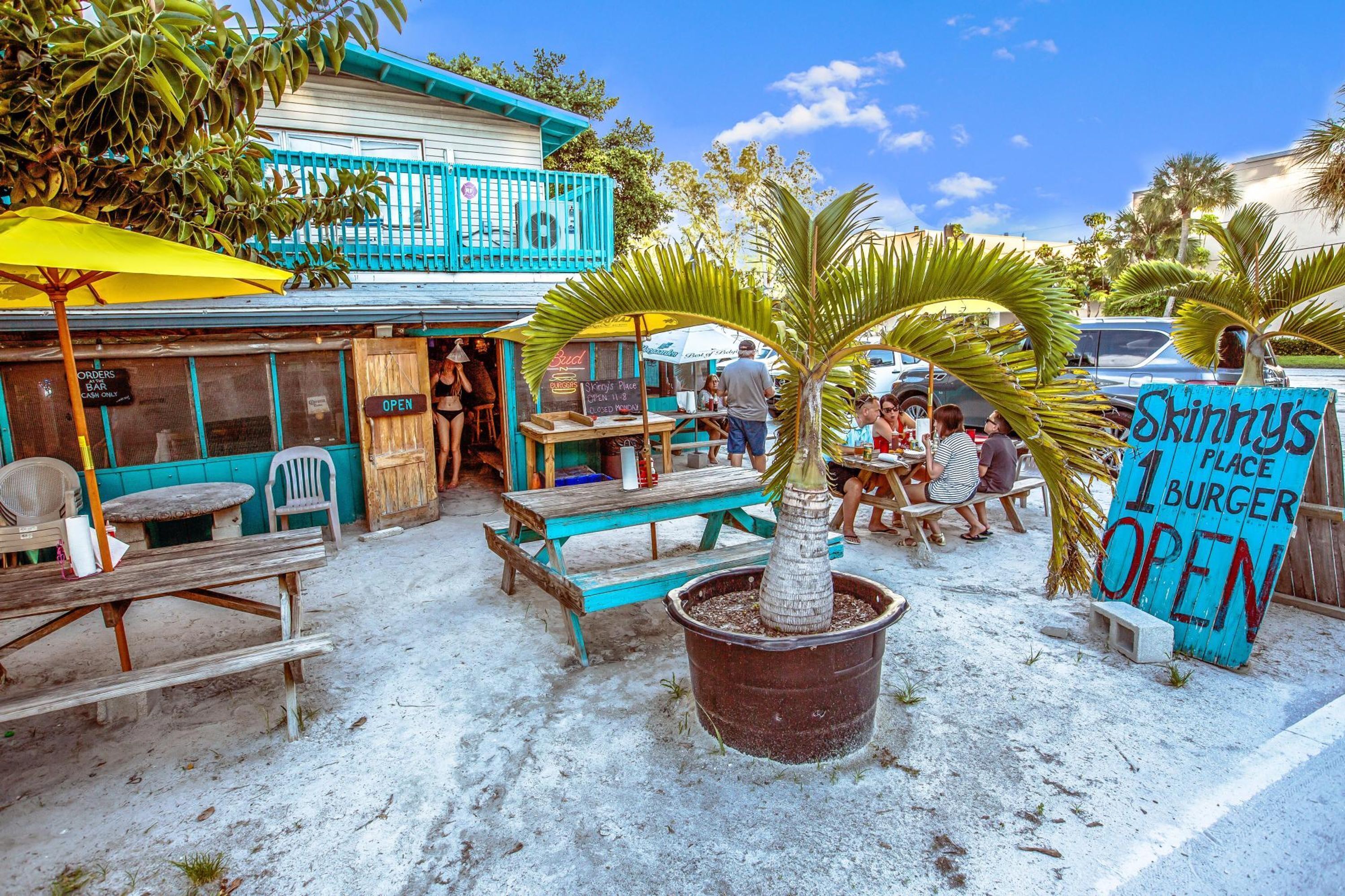 An outdoor dining area with wooden tables and benches, a large potted palm tree, and a sign advertising a burger place.