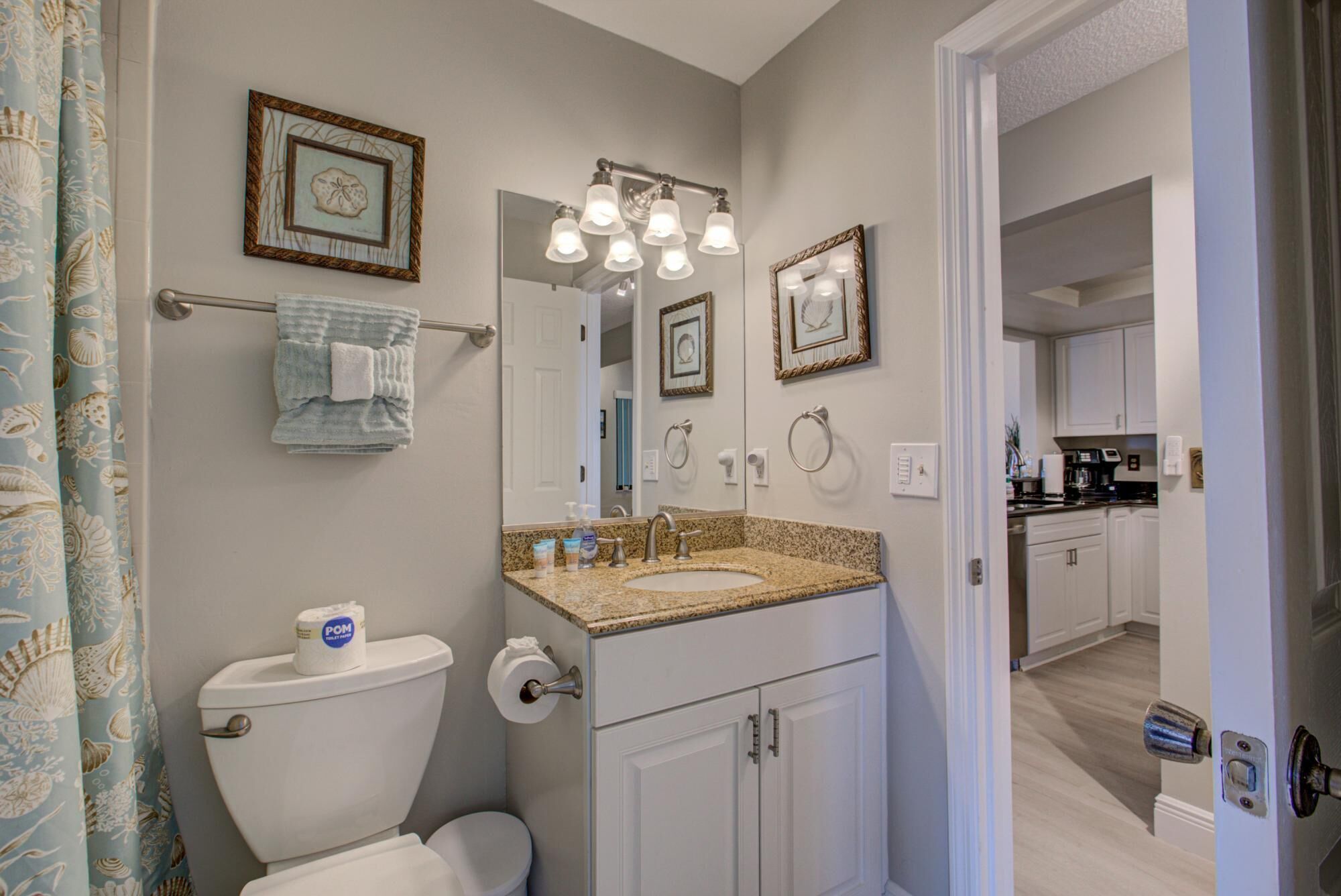 Une salle de bain avec des toilettes, un lavabo et un miroir. Les murs sont ornés de tableaux encadrés, il y a un porte-serviettes et un rideau de douche à motifs de coquillages.