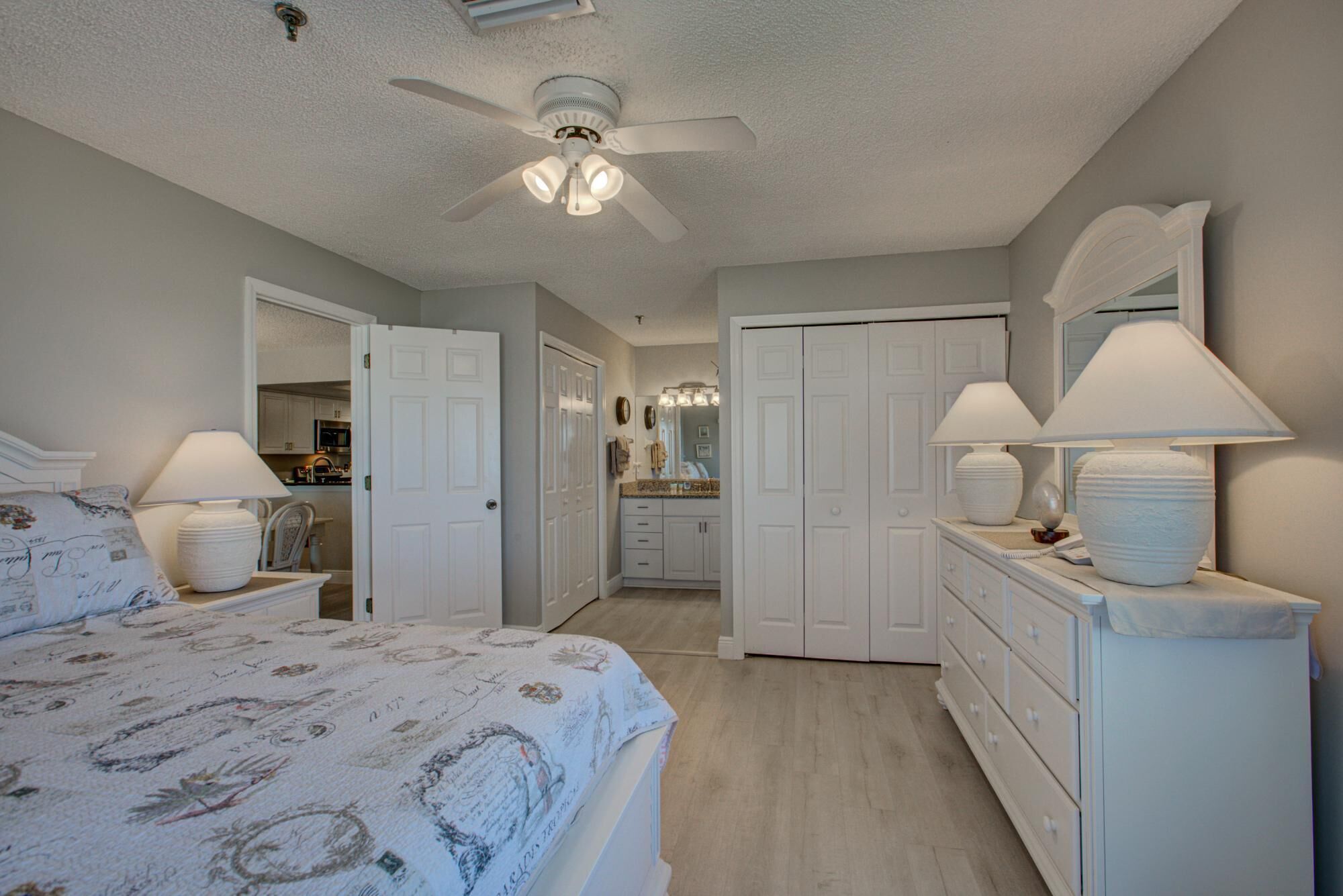A bedroom with a bed, a ceiling fan, a dresser with lamps, and a bathroom visible through an open door.