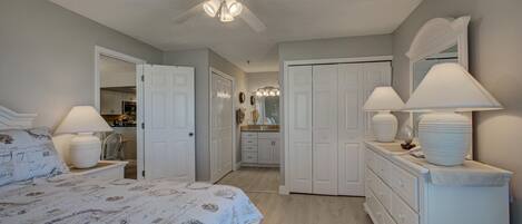 A bedroom with a bed, a ceiling fan, a dresser with lamps, and a bathroom visible through an open door.