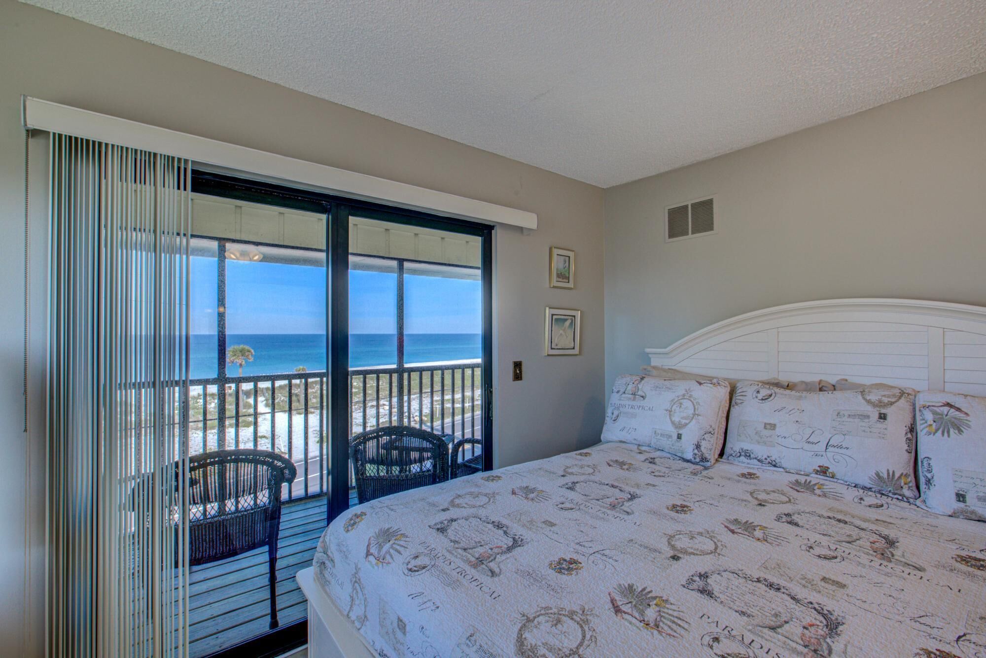 A bedroom with a large bed, a view of the ocean through a sliding door, and a balcony with chairs.