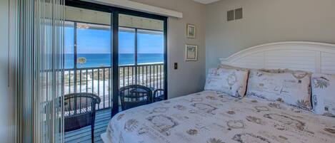 A bedroom with a large bed, a view of the ocean through a sliding door, and a balcony with chairs.