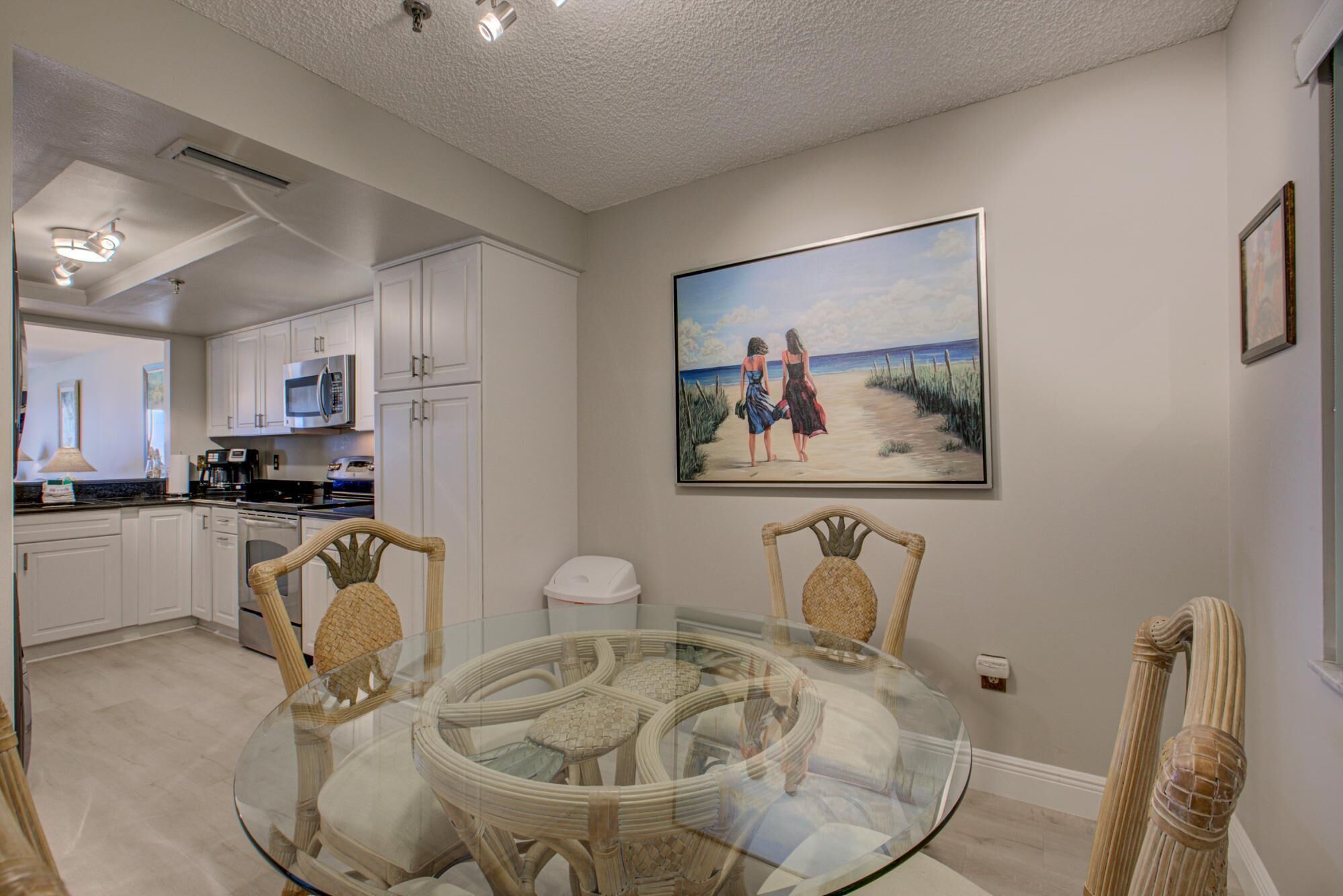 A modern kitchen with white cabinets, a microwave, and a coffee maker. There is a glass dining table with wicker chairs. A painting of two women on a beach is on the wall.