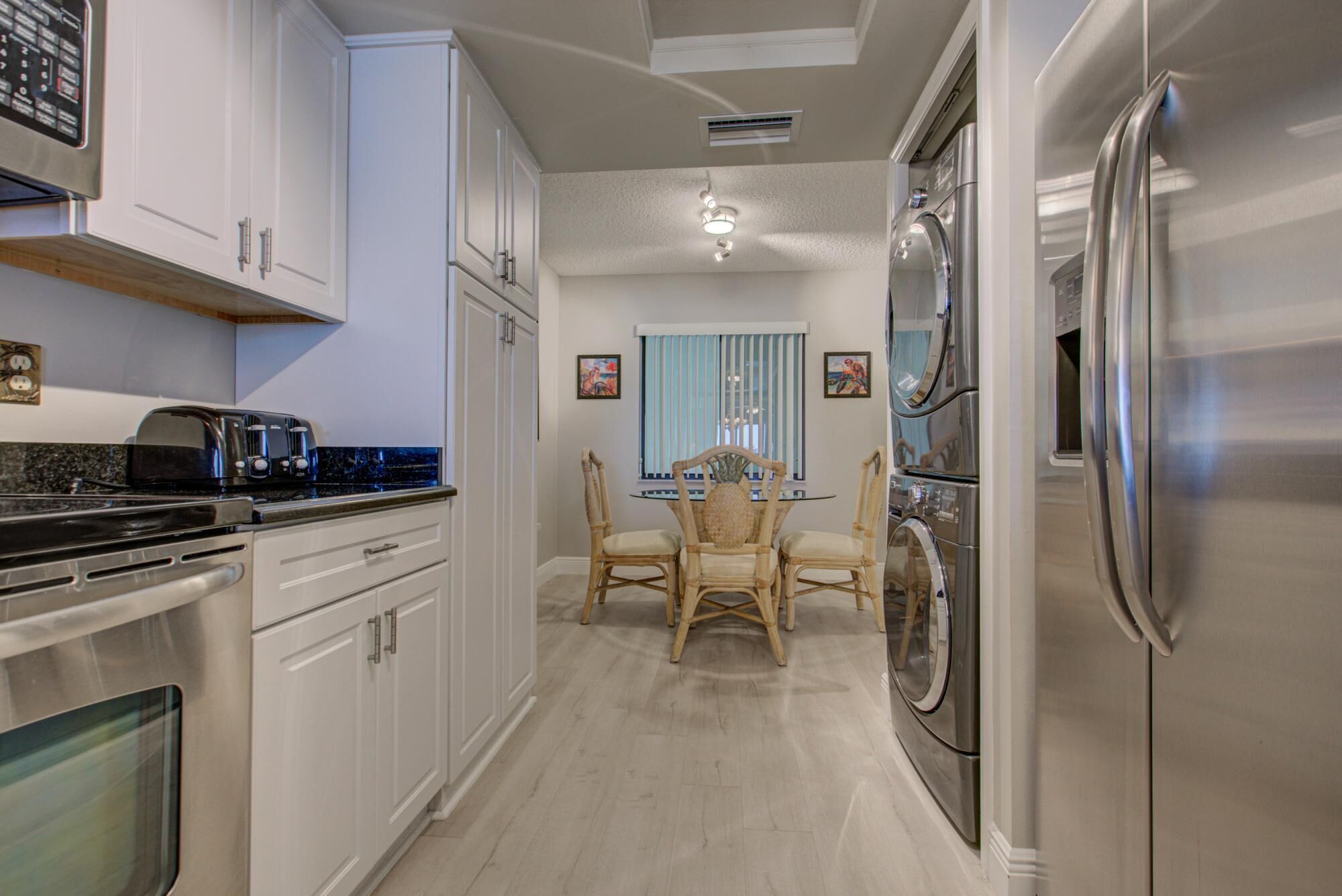 A kitchen with white cabinets, a stainless steel refrigerator, a dining table with chairs, and a window with blinds.