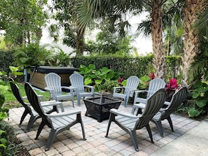 An outdoor seating area with four Adirondack chairs and a square table, surrounded by lush greenery and palm trees.
