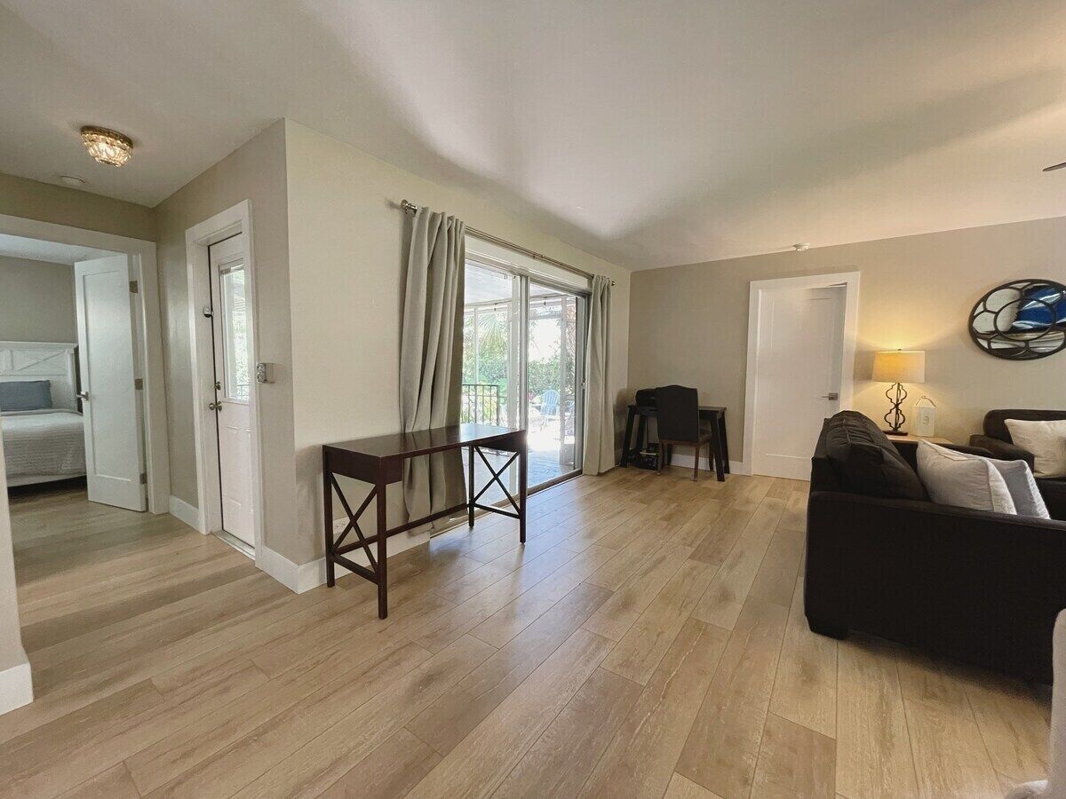 A spacious living area with a wooden floor, a dark-colored sofa, a small table, and a window with curtains.