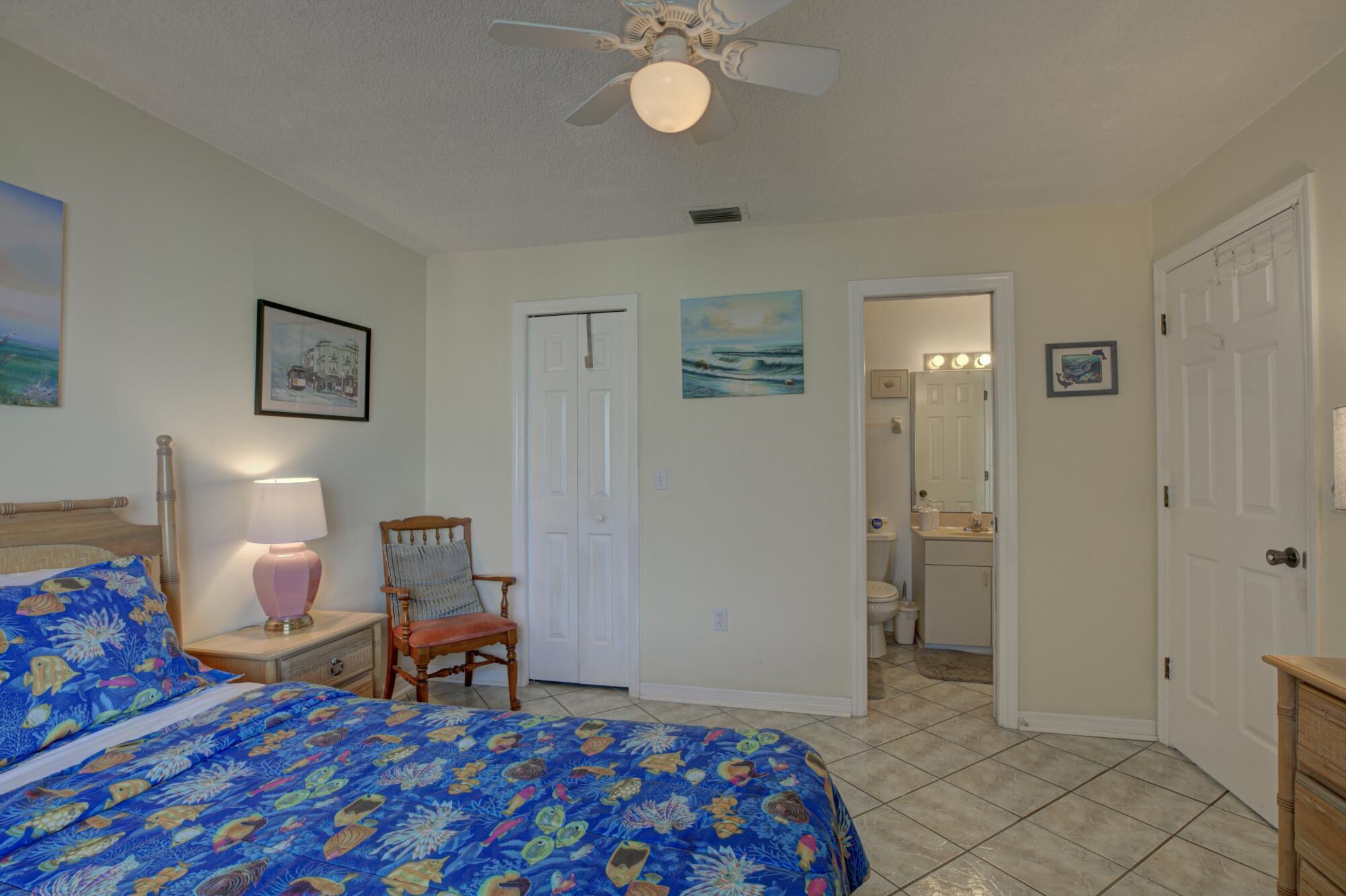 A bedroom with a bed, a chair, a nightstand, a lamp, a ceiling fan, and a bathroom visible through an open door.
