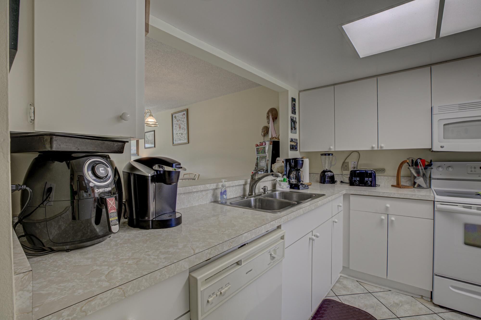 Una cocina compacta con armarios blancos, microondas integrado, cafetera y hervidor.