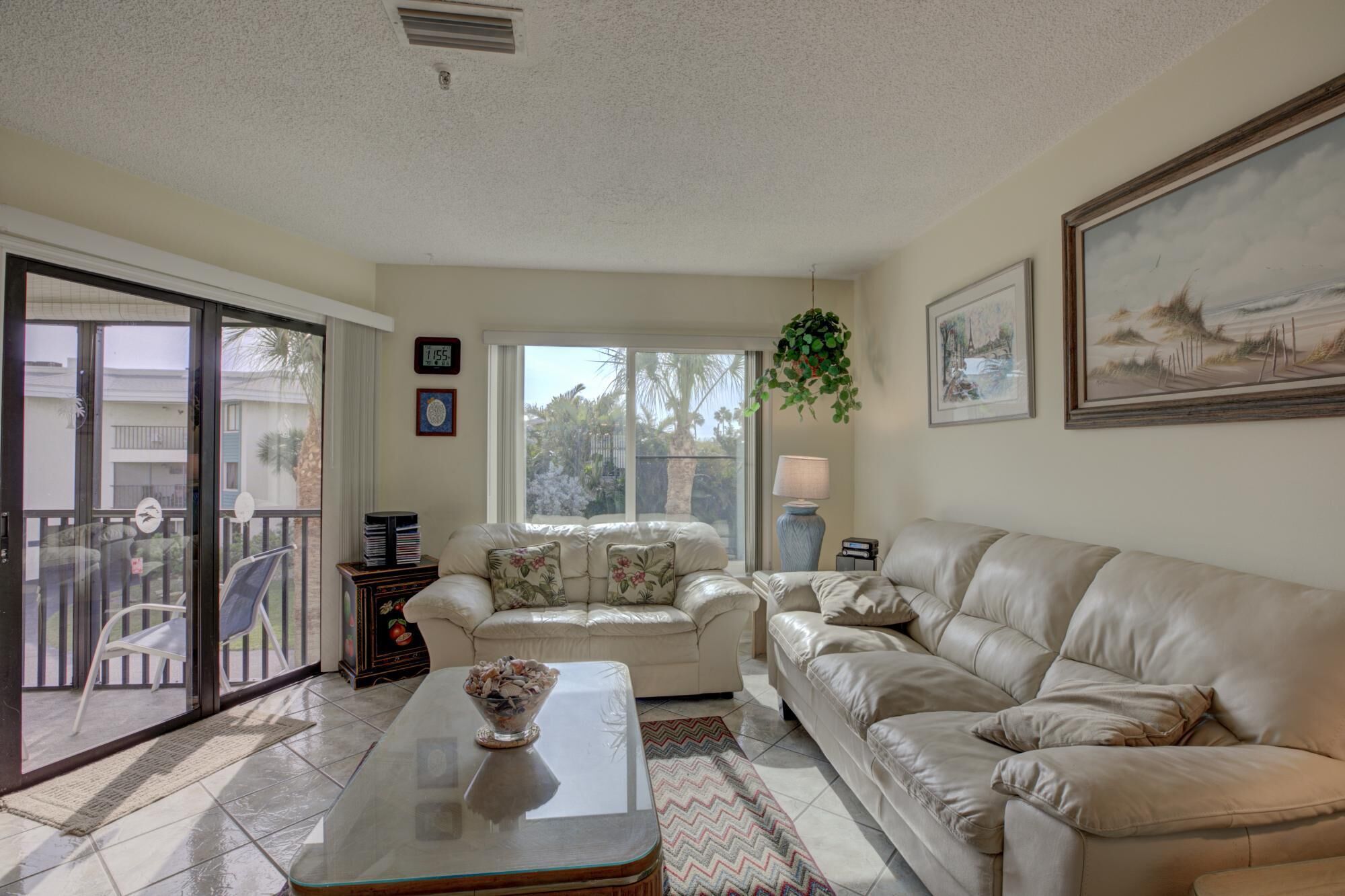 A living room with a sofa, a coffee table, and a TV. There is a window with a view of palm trees and a balcony.