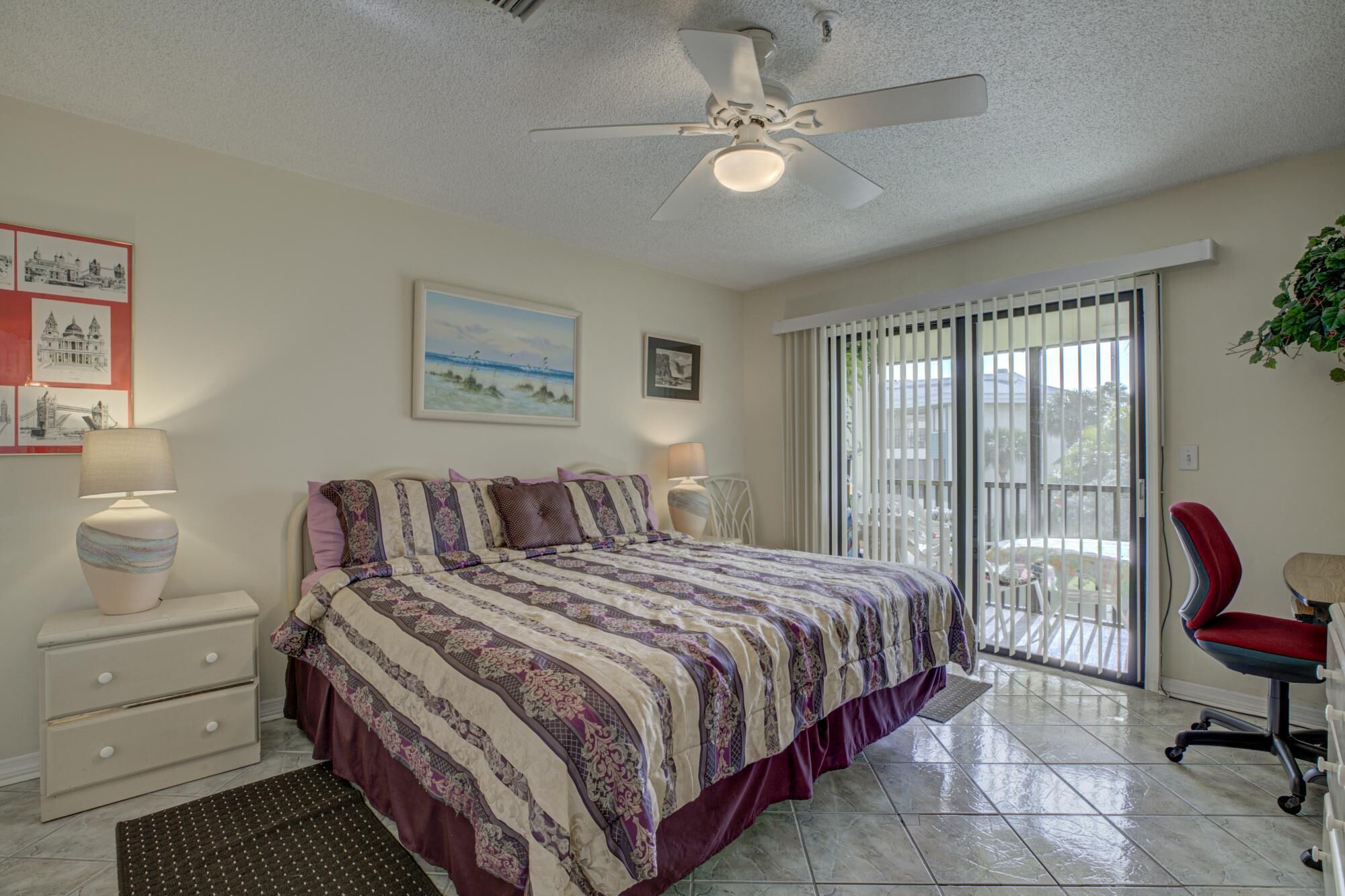A bedroom with a bed, a nightstand, a desk, a chair, a ceiling fan, and a window with blinds.