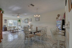 A spacious living room with a dining area, featuring a large window, ceiling fans, and a collection of framed artwork. - Anna Maria Island Beach Bay Breeze (Bradenton Beach)