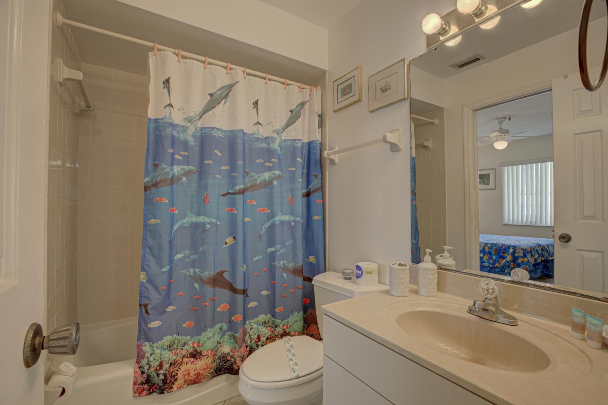 A bathroom with a shower curtain featuring marine life, a toilet, a sink with toiletries, and a mirror.