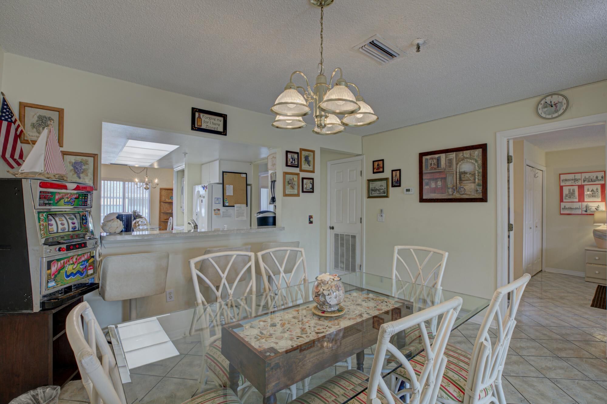 A dining area with a glass table, white chairs, and a chandelier. There is a refrigerator with a slot for a game machine, and a door leading to another room.