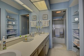 Ein Badezimmer mit einem großen Spiegel, zwei Waschbecken, einer Dusche und Regalen mit Handtüchern und Toilettenartikeln.