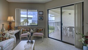 A cozy living room with a sofa, a coffee table, and a lamp. There is a large window with a view of a house and palm trees. A sliding glass door leads to an outdoor patio with chairs and a table. - Anna Maria Island Bridge Port Beach Resort (Bradenton Beach)