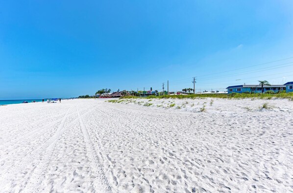 Une vaste plage de sable avec des empreintes, un ciel bleu dégagé et des bâtiments en arrière-plan.