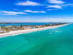 Une vue aérienne d’une station balnéaire dotée d’une longue plage, d’eaux turquoise et limpides, et d’un ensemble dense de bâtiments le long du littoral.
